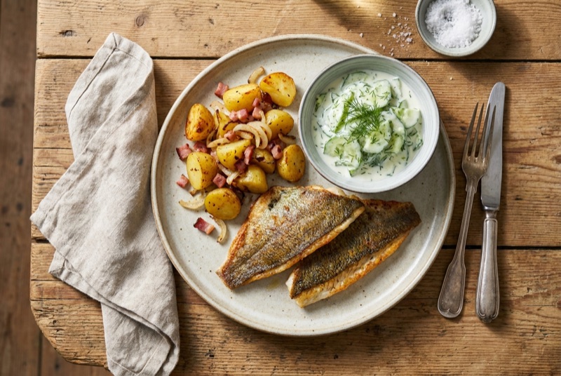 Gebratener Fisch mit Bratkartoffeln und Gurkensalat auf Holztisch